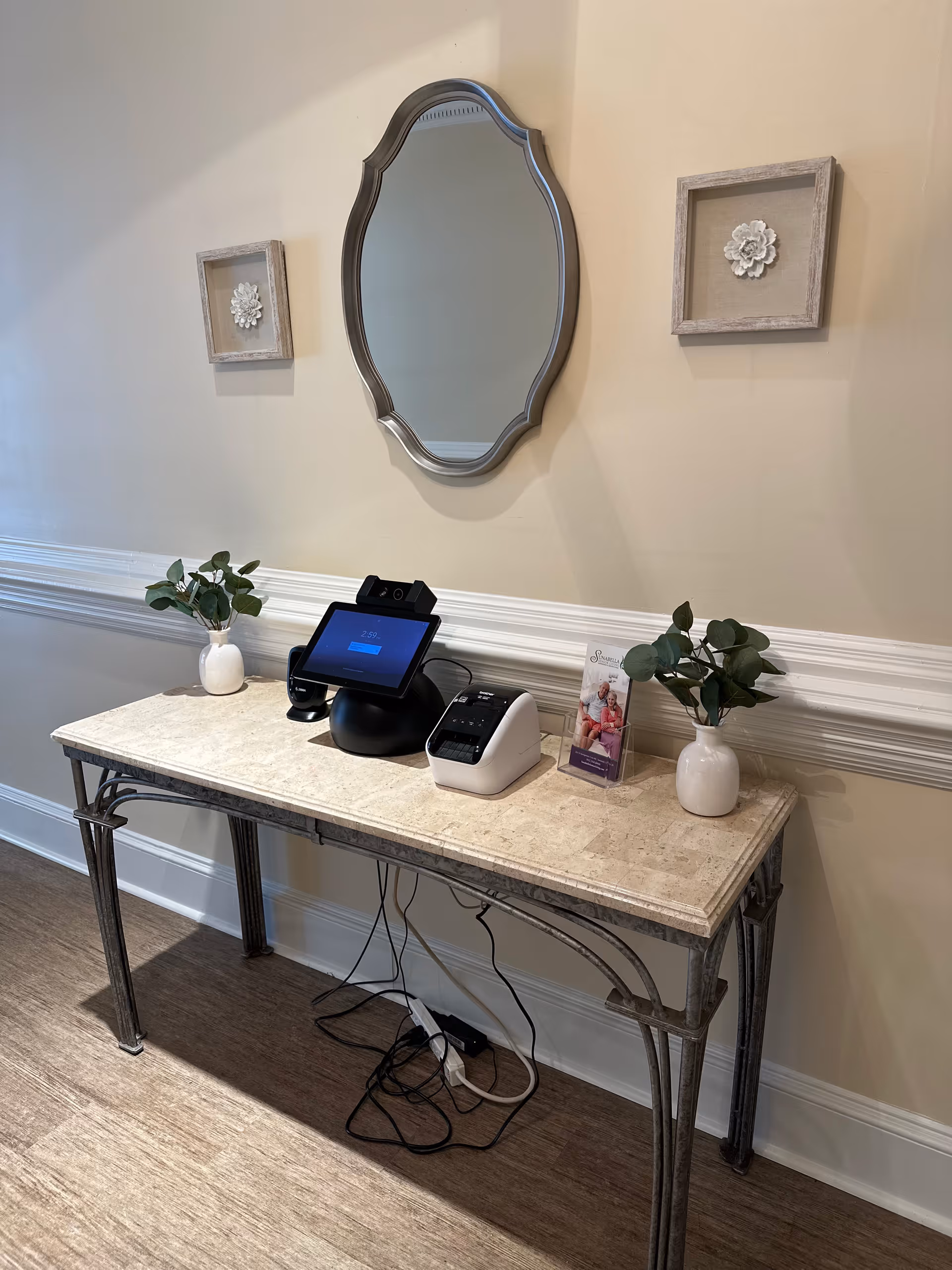 A decorative console table with a marble top and metal legs against a beige wall. On the table are two small white vases with green leafy plants, a digital device with a screen, a small white printer, and a brochure holder with a brochure. Above the table is an ornate oval mirror flanked by two square wall decorations with floral designs.