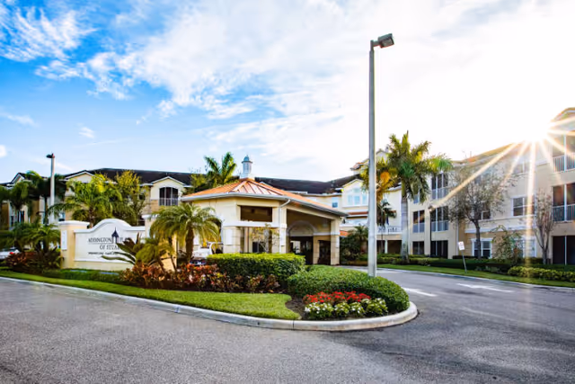 Exterior view of Addington Place of Stuart, showing a multi-story building with balconies, palm trees, landscaped greenery, and a driveway with a covered entrance under a partly cloudy sky with the sun shining brightly.