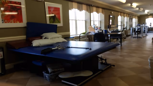 A rehabilitation therapy room with a large blue treatment table, pillows, walkers and exercise equipment lined along windows in a long, well-lit space.