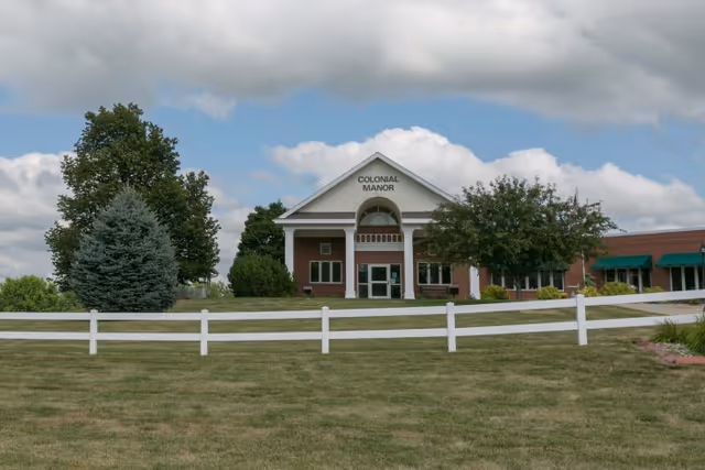 Front exterior view of Colonial Manor building with a white fence in the foreground, green trees, and a partly cloudy sky.