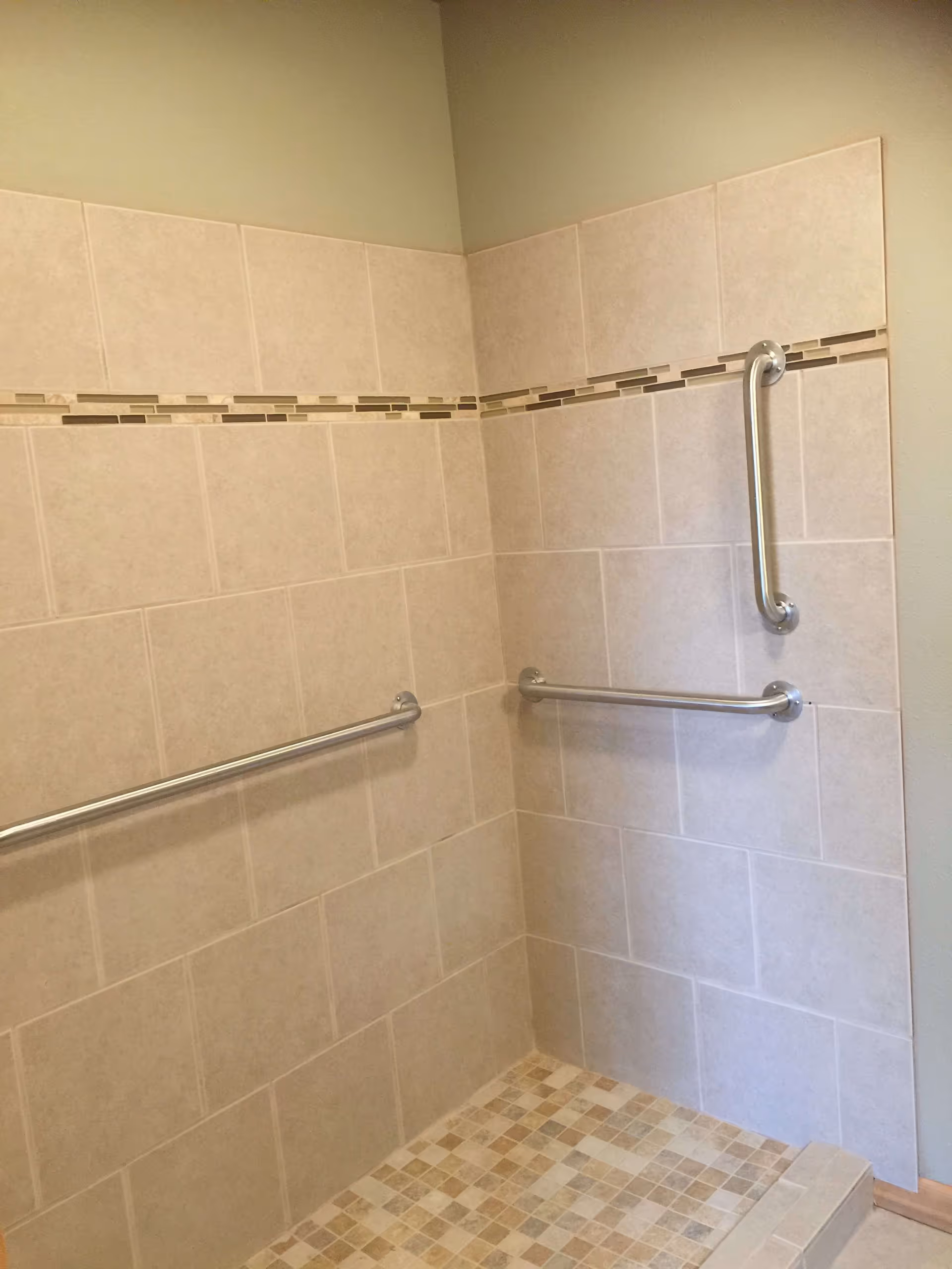 A tiled shower area with beige and light brown tiles, featuring two horizontal grab bars and one vertical grab bar for support. The shower floor has small square mosaic tiles in various shades of beige and brown.