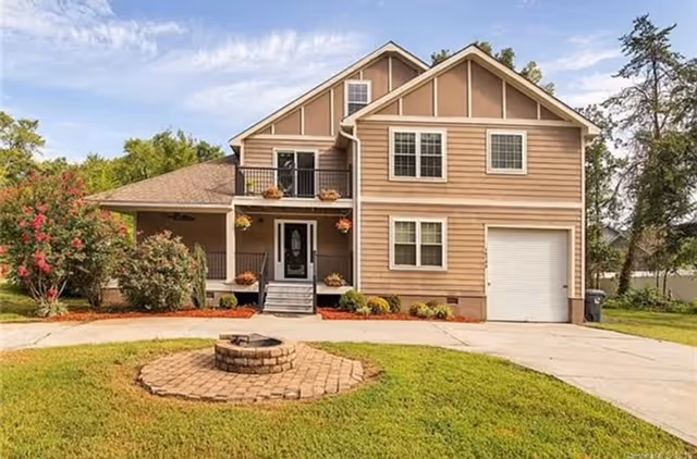 A two-story beige house with a circular brick fire pit in the front yard and a driveway leading to a garage.