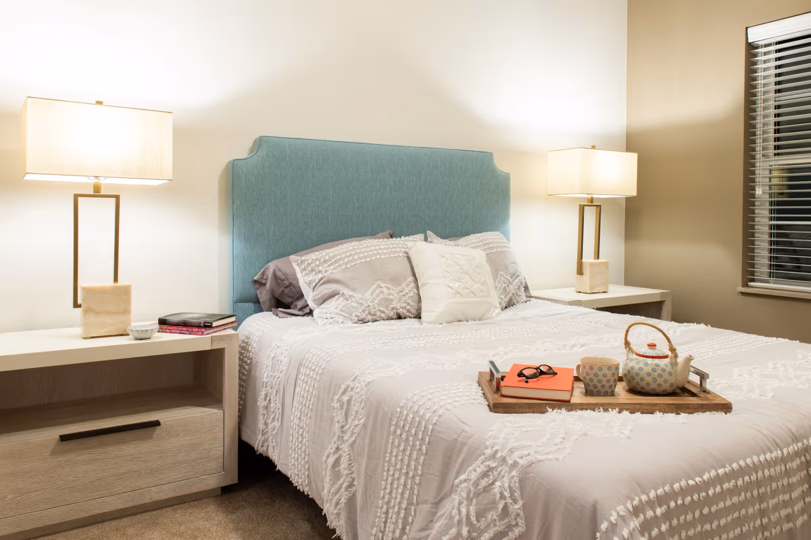 A neatly made bed with a light blue upholstered headboard and white textured bedding. There are two bedside tables, each with a modern lamp. On the bed is a wooden tray holding a teapot, a cup, a pair of glasses, and a book. A window with blinds is visible on the right side of the room.