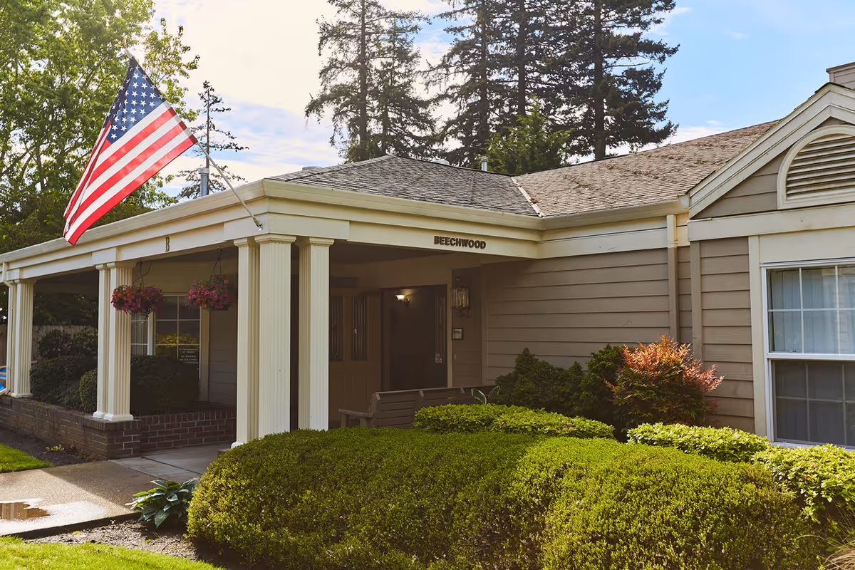 Exterior view of a senior living facility building named Beechwood with beige siding, white columns, hanging flower baskets, an American flag, and well-maintained green bushes and trees.