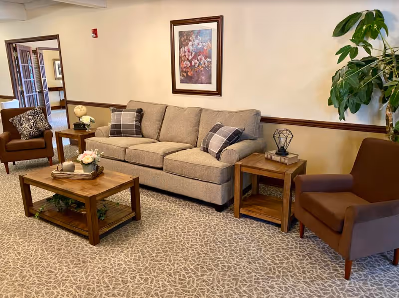 A cozy seating area with a beige sofa, two brown armchairs, wooden coffee and side tables, framed artwork on the wall, and a potted plant.