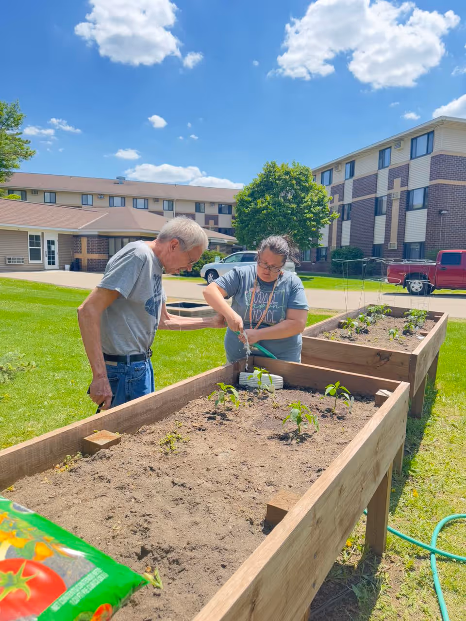 Two people tending and watering raised garden beds on the lawn outside an assisted living building under a blue sky.