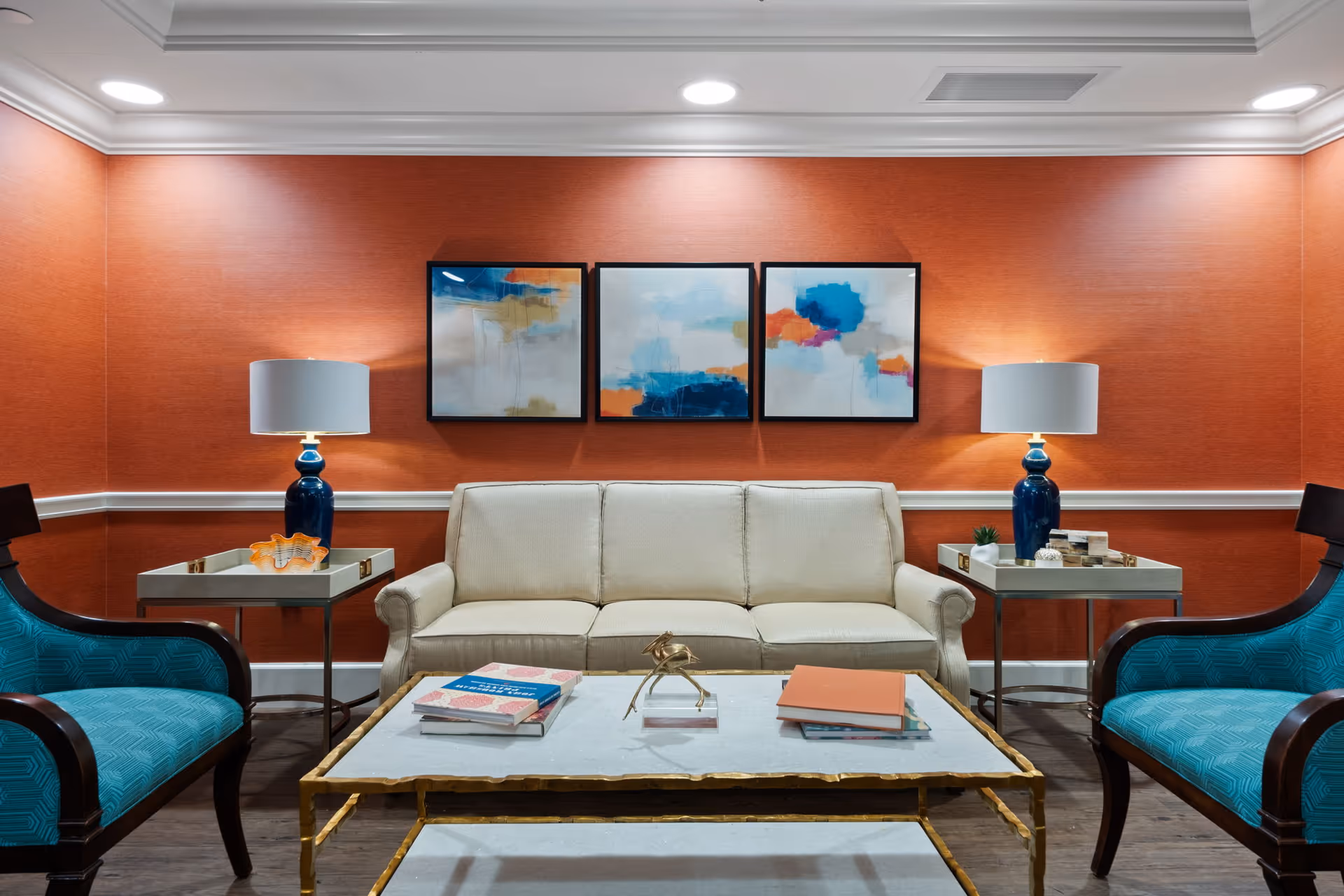 A cozy living room with a beige sofa centered against a burnt orange wall. Above the sofa are three abstract paintings in shades of blue, orange, and white. On either side of the sofa are side tables with blue lamps and decorative items. Two teal upholstered armchairs with dark wooden frames face the sofa. A marble-top coffee table with gold trim holds books and a small decorative sculpture.