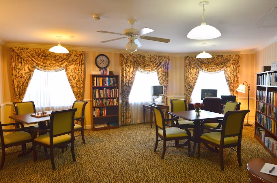 A cozy common room with two square tables surrounded by green cushioned chairs. The room has floral curtains on three windows, a ceiling fan, and bookshelves filled with books. There are two computer desks with monitors near the windows and a floor lamp in the corner.