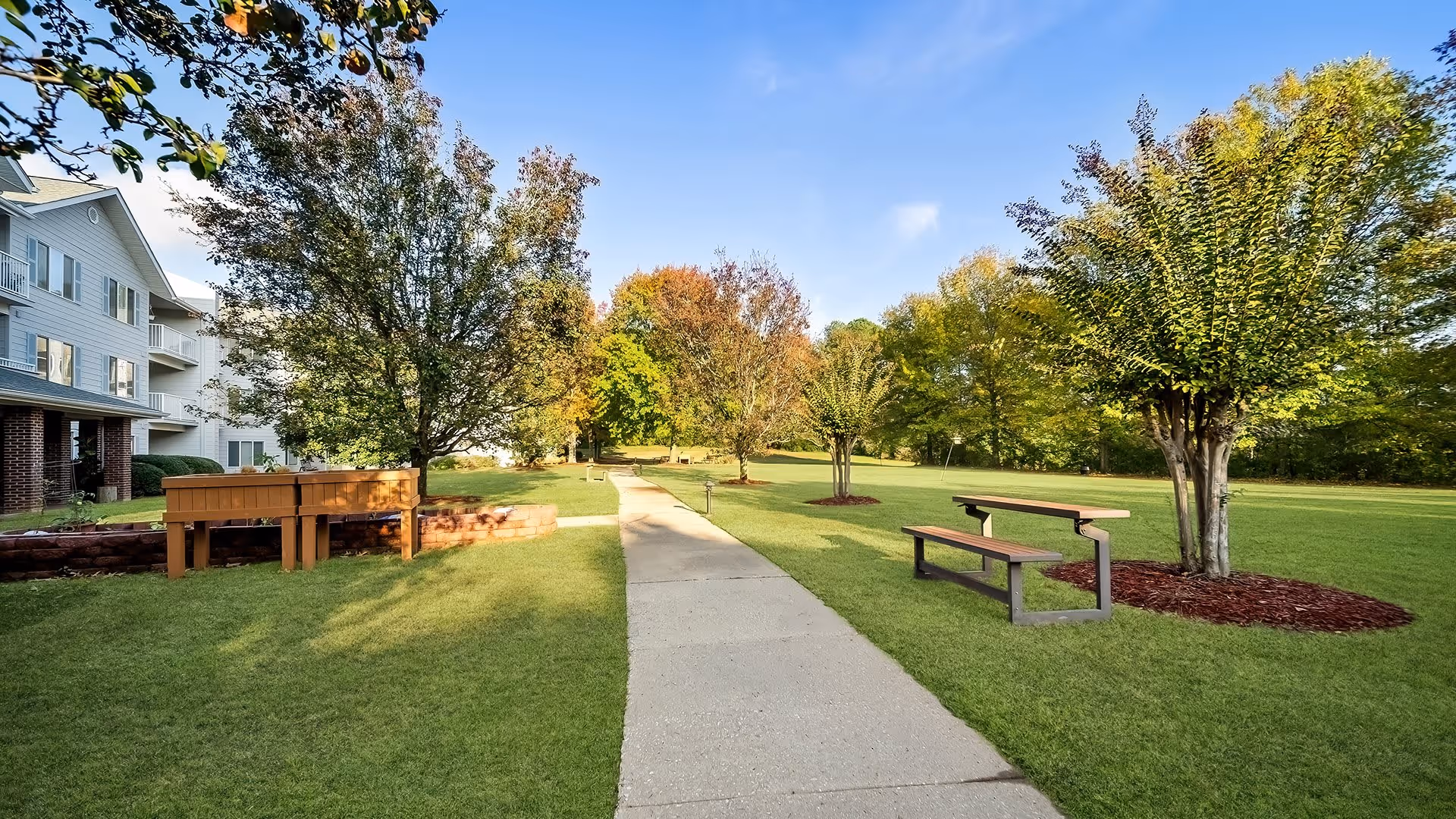A paved walkway runs through a green lawn with several trees and benches on either side. To the left, part of a multi-story building with balconies is visible. The sky is clear and blue, suggesting a sunny day.