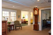 Interior view of a senior living facility showing a hallway with beige carpet and walls. There is a wooden grandfather clock against the wall, a green upholstered chair next to a small wooden table with a plant, and a glimpse into adjacent rooms with more seating and tables. The space is well-lit with natural light coming through windows and has a calm, welcoming atmosphere.