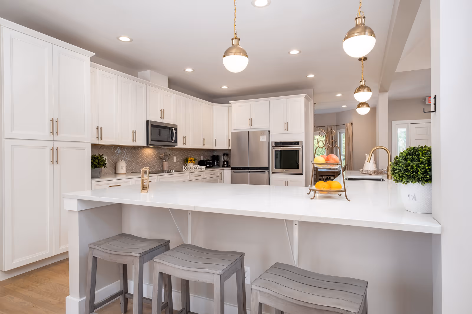 Bright modern kitchen with white cabinetry, a large white island countertop with three gray wooden stools, stainless steel refrigerator and oven, a microwave above the stove, pendant lights hanging from the ceiling, and a small plant and fruit stand on the island.