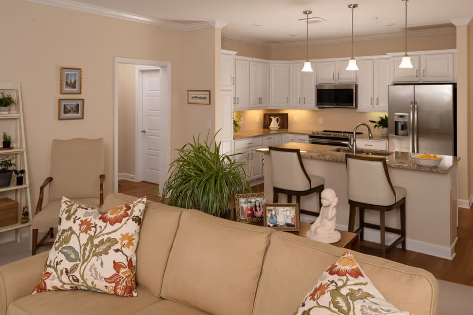 A cozy and well-lit interior space showing a beige sofa with floral patterned pillows in the foreground, a beige armchair, a green potted plant, and a white kitchen with granite countertops, stainless steel appliances, and two bar stools. Family photos and a small statue are displayed on a wooden table behind the sofa.