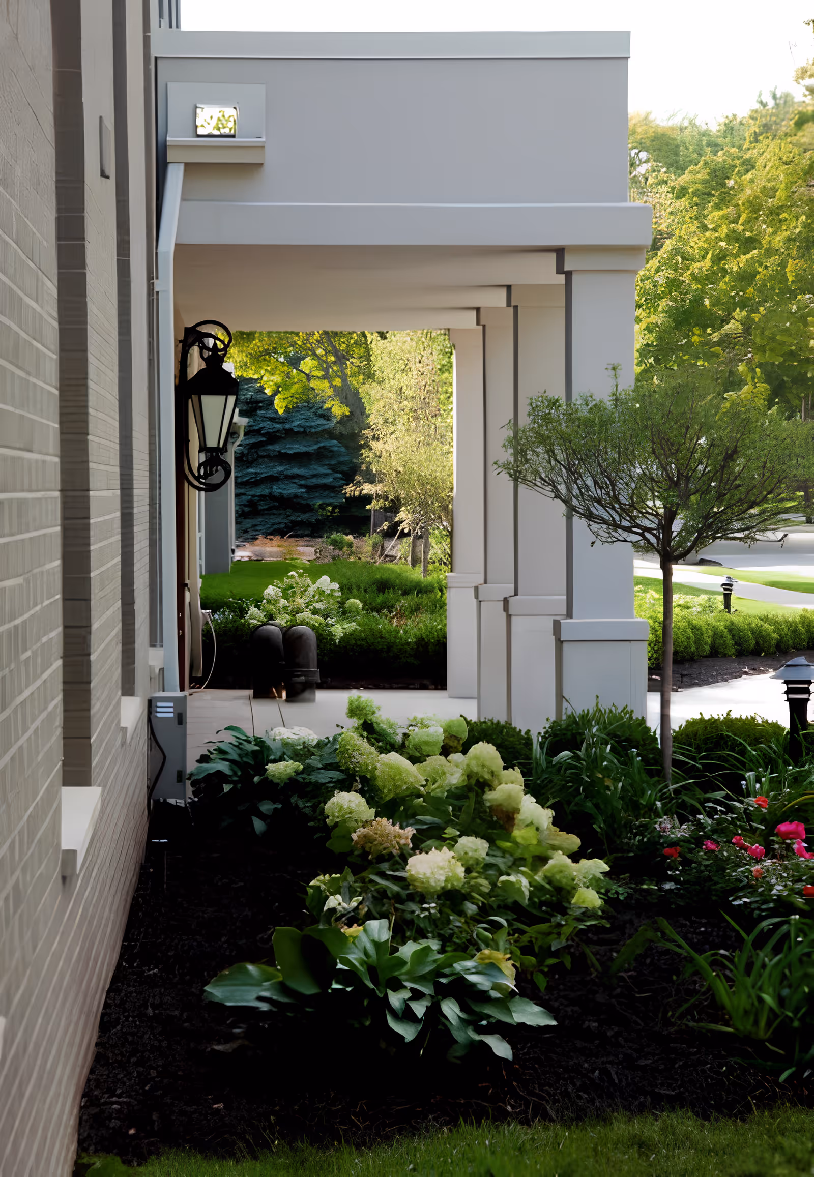 Covered exterior walkway beside the building with white columns, a hanging lantern, and landscaped flower beds.