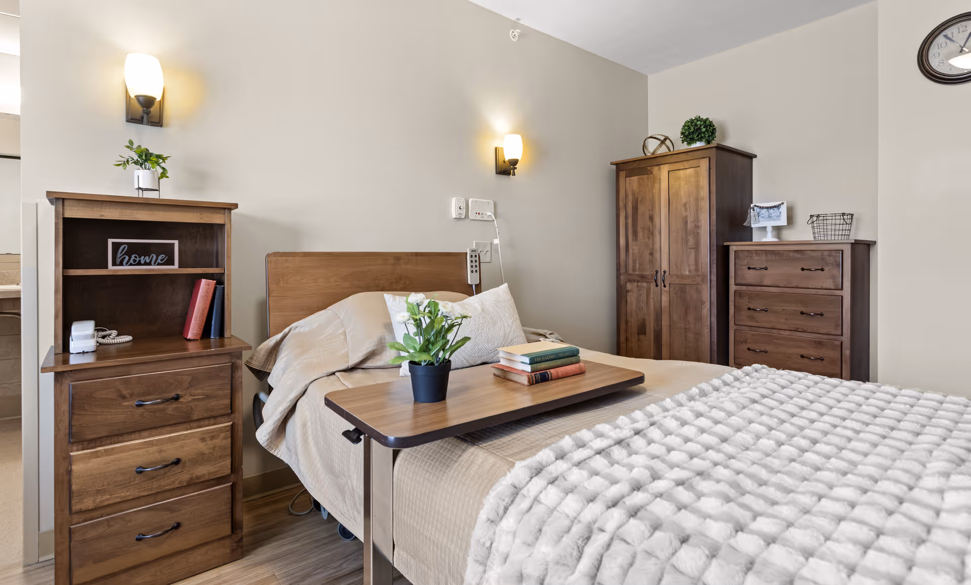 A cozy bedroom in a senior living facility featuring a single bed with beige bedding and a textured white blanket. A wooden overbed table holds a small potted plant and a stack of books. The room includes wooden furniture such as a nightstand with drawers, a wardrobe, and a chest of drawers. Two wall-mounted lights provide warm lighting, and a clock is visible on the wall.