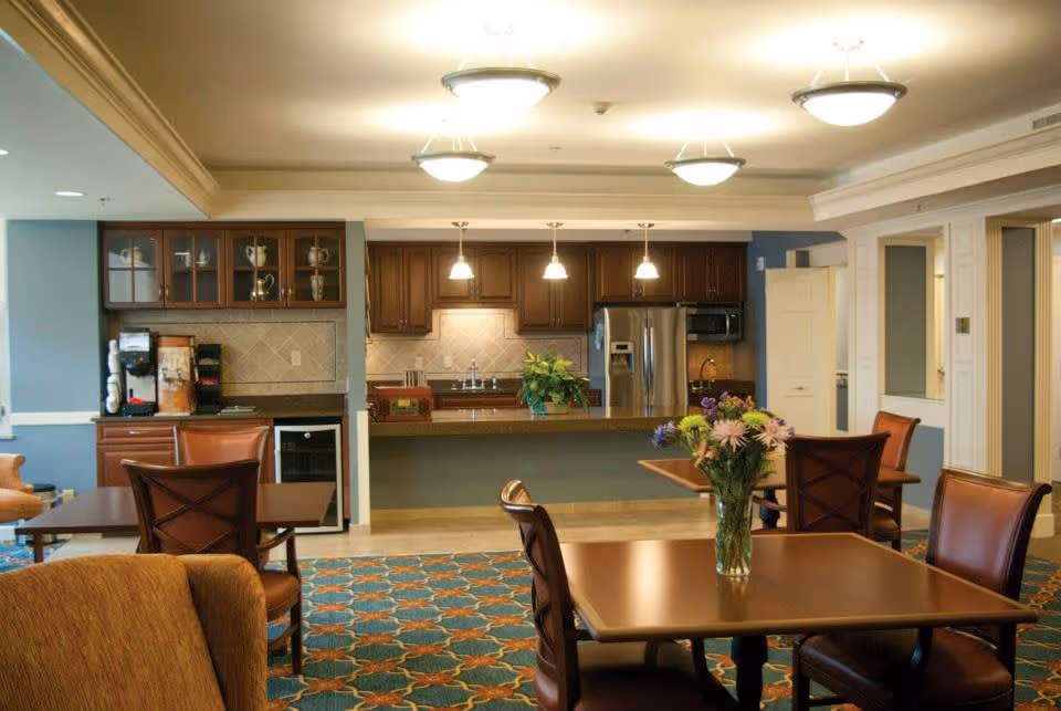Interior view of a senior living facility dining area with wooden tables and chairs, a vase with flowers on one table, and a kitchen area in the background featuring wooden cabinets, a stainless steel refrigerator, and pendant lights.