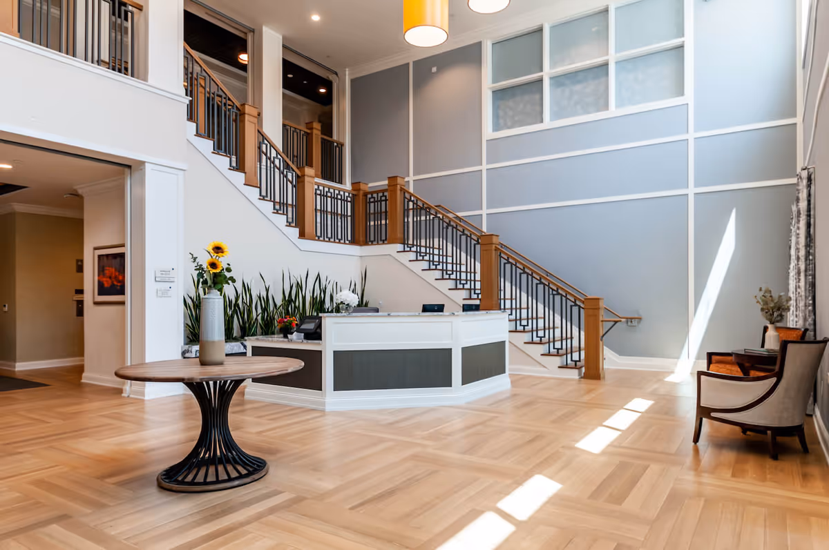 Bright staffed lobby with a reception desk, central round table with flowers, staircase, and seating area.