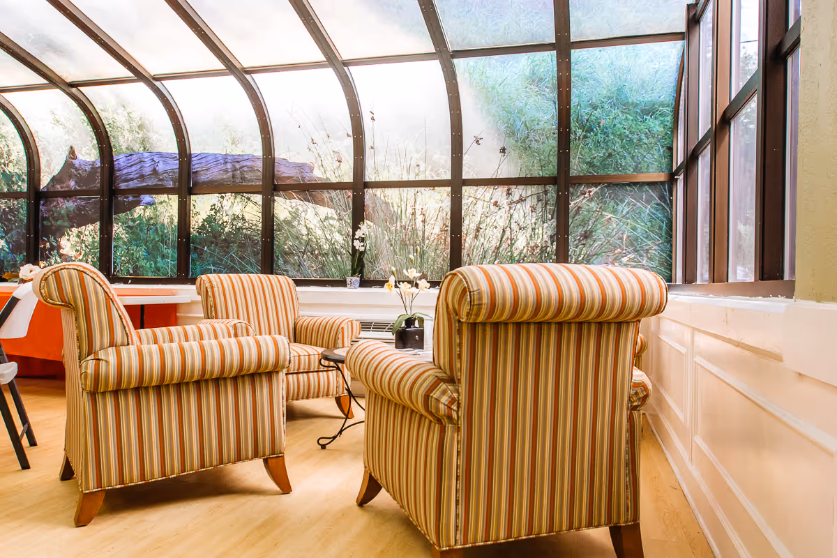 Sunlit seating area with three striped armchairs around a small table in front of a large curved glass window showing greenery.