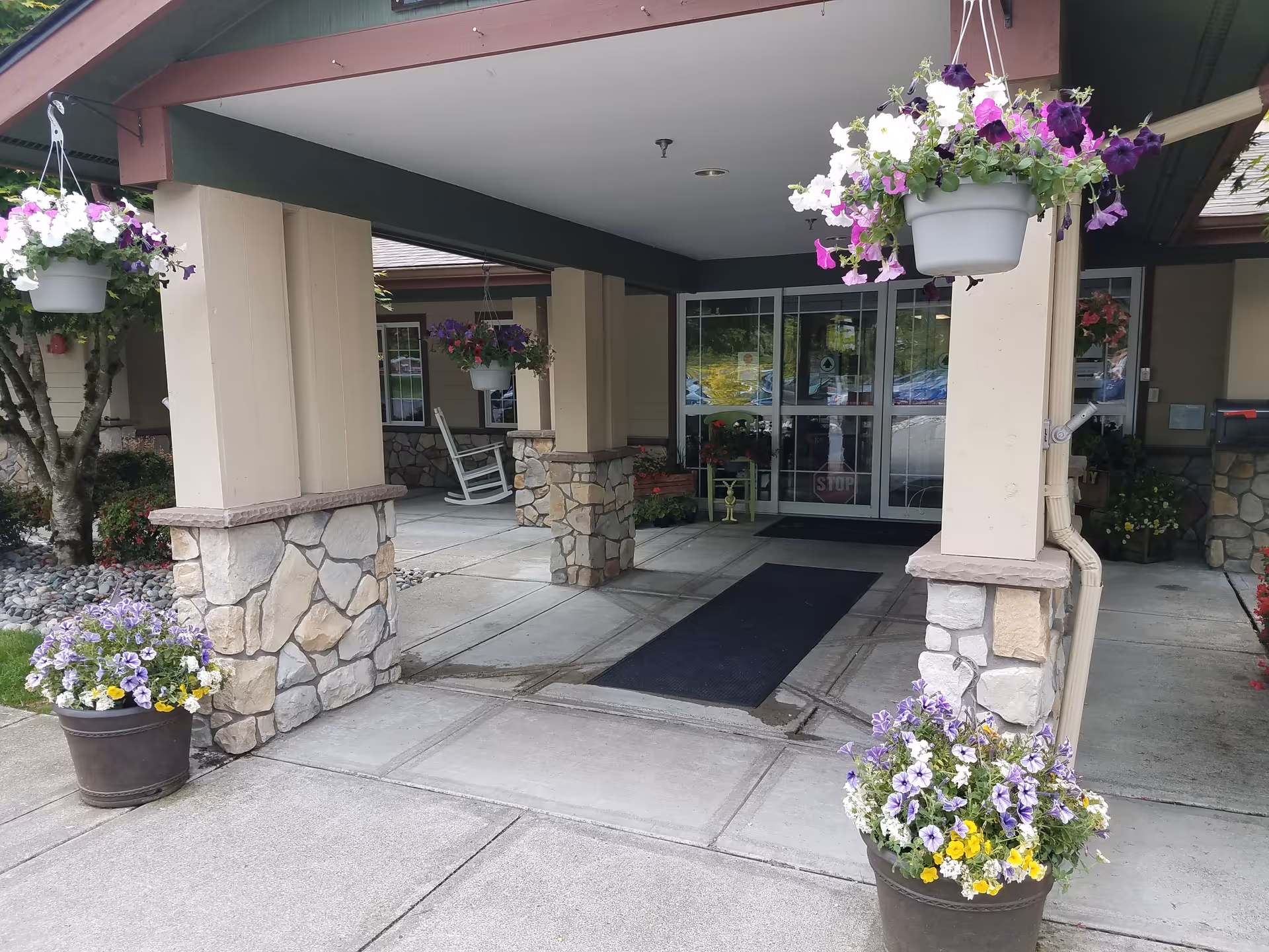 Covered entrance to Brookdale Arbor Place with stone columns, hanging flower baskets, potted flowers, and glass double doors.