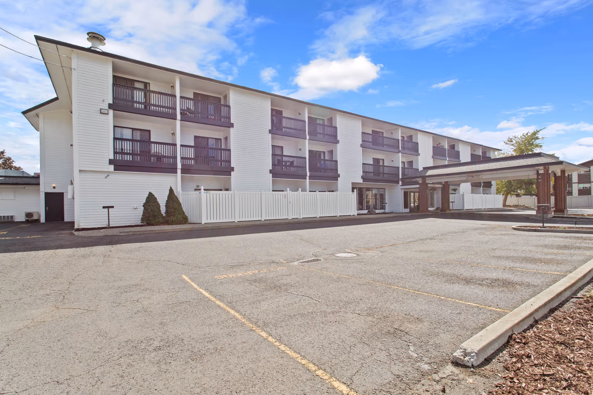 Exterior view of a three-story senior living facility building with white siding and dark brown balconies. The building has a covered entrance area and a large empty parking lot in front. The sky is partly cloudy with blue patches.