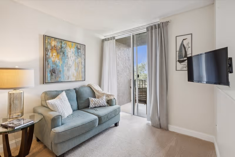 A cozy living room with a light blue sofa adorned with three decorative pillows. A glass side table with a lamp and a book sits next to the sofa. Above the sofa hangs an abstract painting with blue, yellow, and white tones. To the right, there is a wall-mounted flat-screen TV and a framed artwork of a sailboat. Sliding glass doors with curtains lead to a small outdoor balcony with a chair visible.