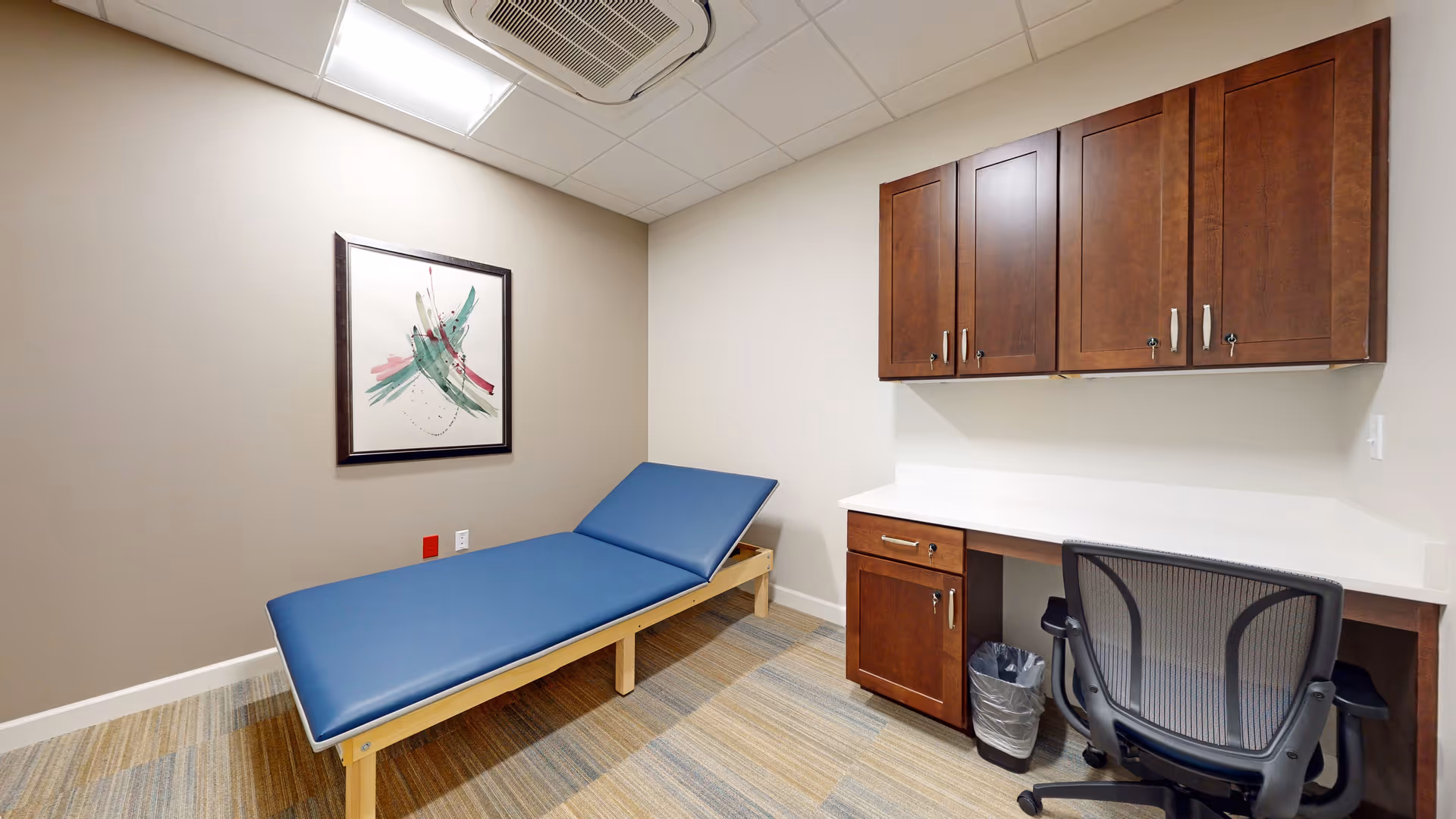 Small clinical-style room with a blue examination table, wooden cabinets, and a desk with an office chair.