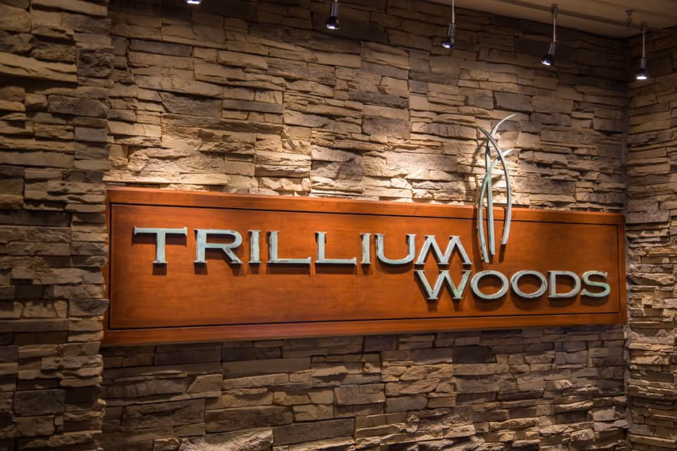 A wooden sign mounted on a stone wall with the text 'TRILLIUM WOODS' and a decorative plant-like design above the word 'WOODS'. The sign is illuminated by several spotlights from above.