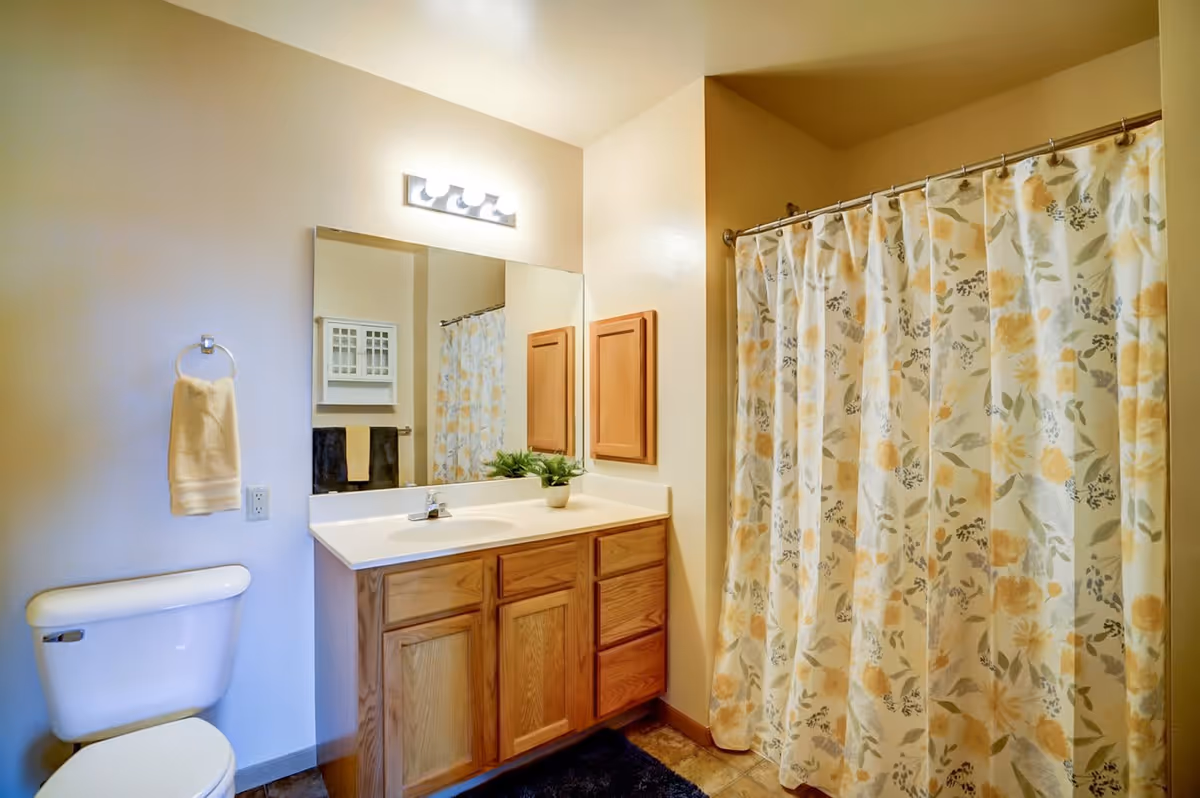 A bathroom with a white toilet, a wooden vanity with a white countertop and sink, a large mirror above the vanity, a towel ring with a beige towel, a floral shower curtain with yellow and green patterns, and a small potted plant on the countertop.