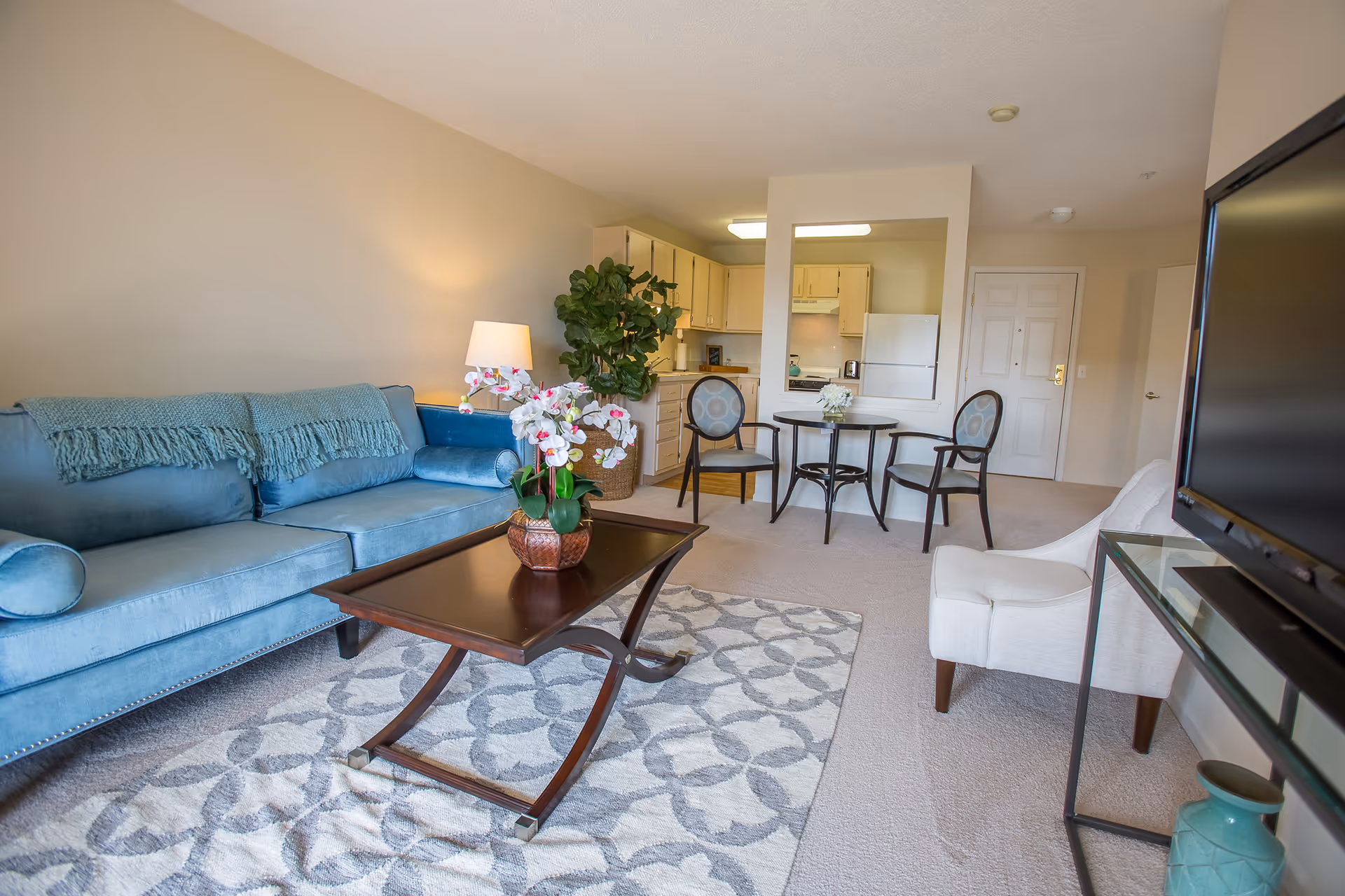 A bright and cozy living room with a blue sofa, a white armchair, a wooden coffee table with a potted orchid, and a patterned rug. In the background, there is a small dining area with a round table and three chairs, and a kitchen with light wood cabinets and white appliances.