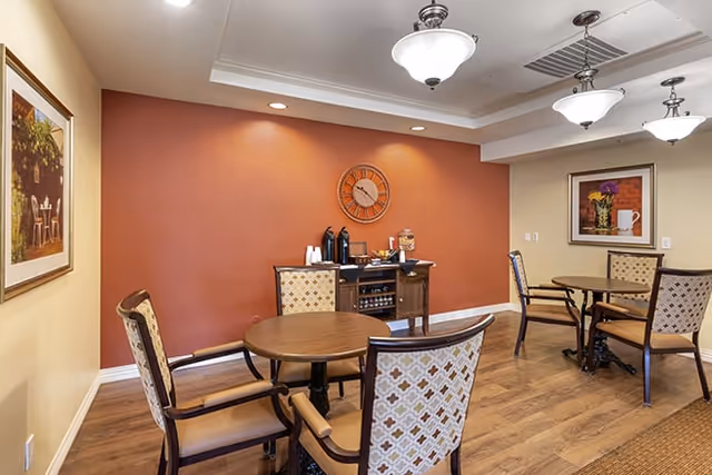 Communal dining room with round wooden tables, patterned upholstered chairs, and a beverage station against a terracotta accent wall.