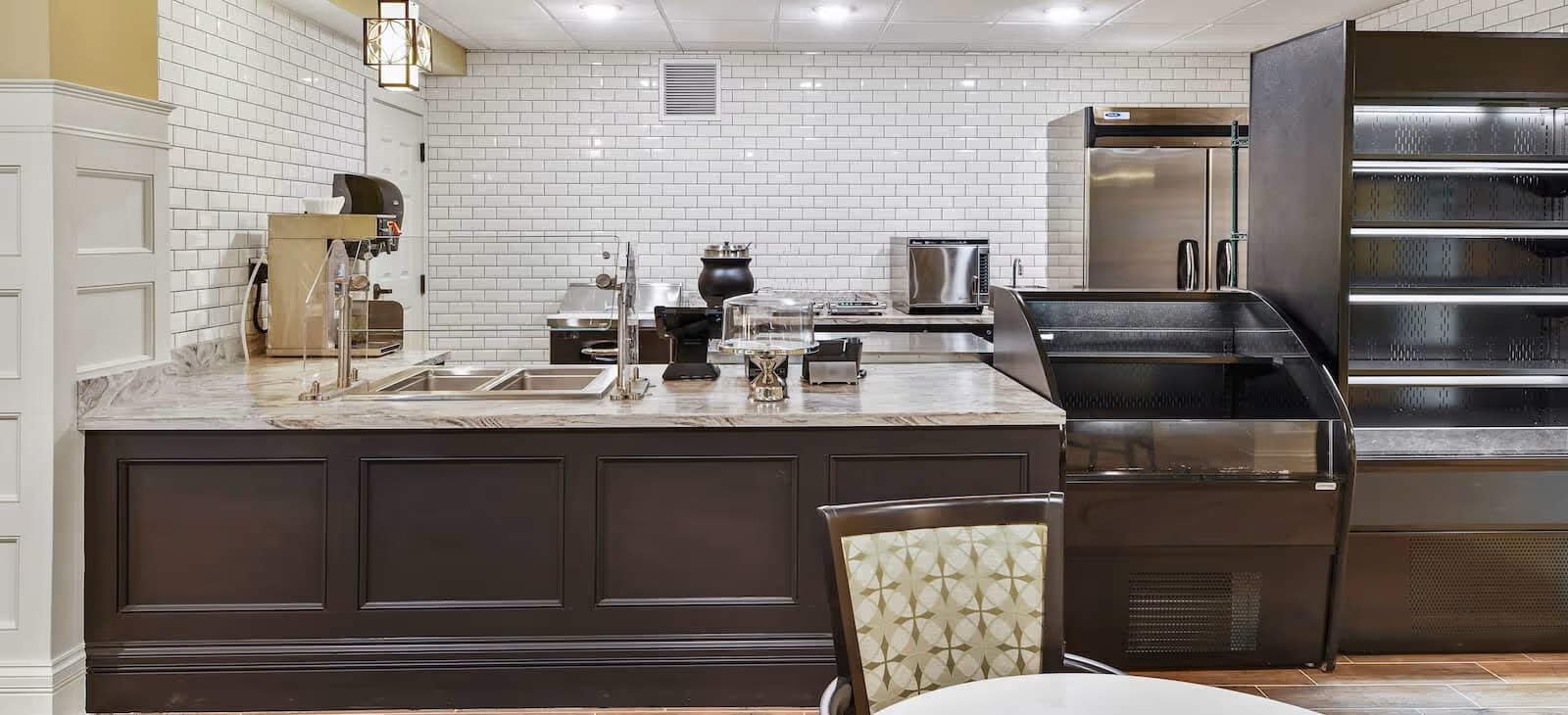 Interior view of a clean, modern kitchen area with a marble countertop, a sink, coffee machine, toaster, and stainless steel refrigerator. There is a display case and shelving unit on the right side, and a chair with a patterned cushion in the foreground.