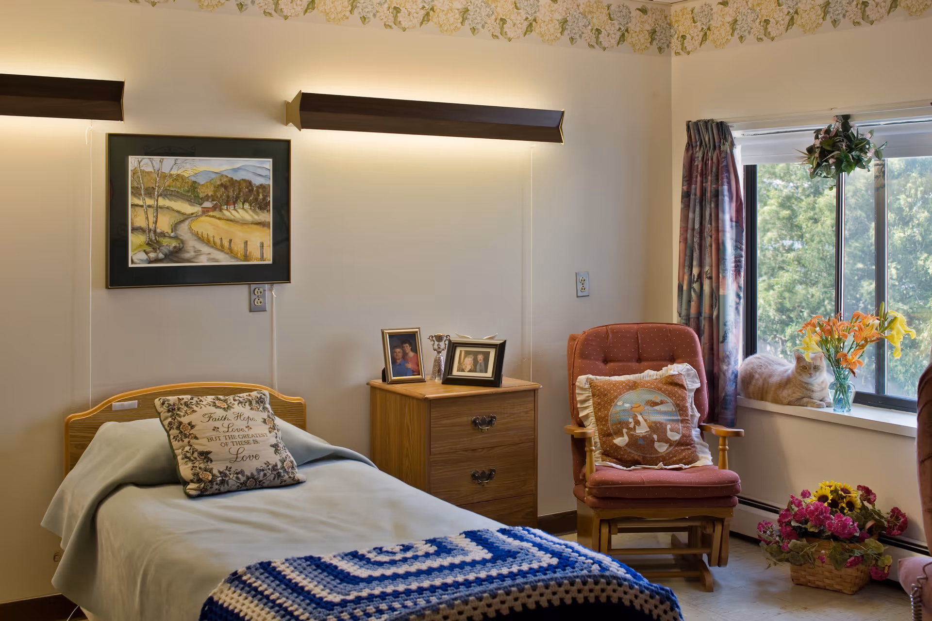 A cozy bedroom in a senior living facility with a single bed covered with a light blue blanket and a decorative pillow. Next to the bed is a wooden nightstand with framed photos and a small trophy. A cushioned armchair with a decorative pillow sits near a window with floral curtains. A cat is resting on the windowsill next to a vase of orange and yellow flowers. There is a basket of flowers on the floor and a landscape painting on the wall above the bed.