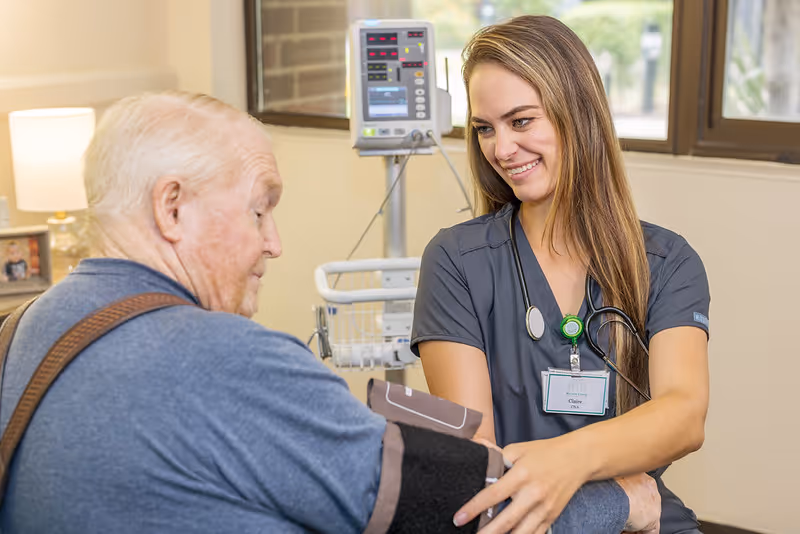 A female healthcare professional wearing scrubs and a stethoscope is smiling while taking the blood pressure of an elderly man in a medical setting with a blood pressure monitor and window in the background.