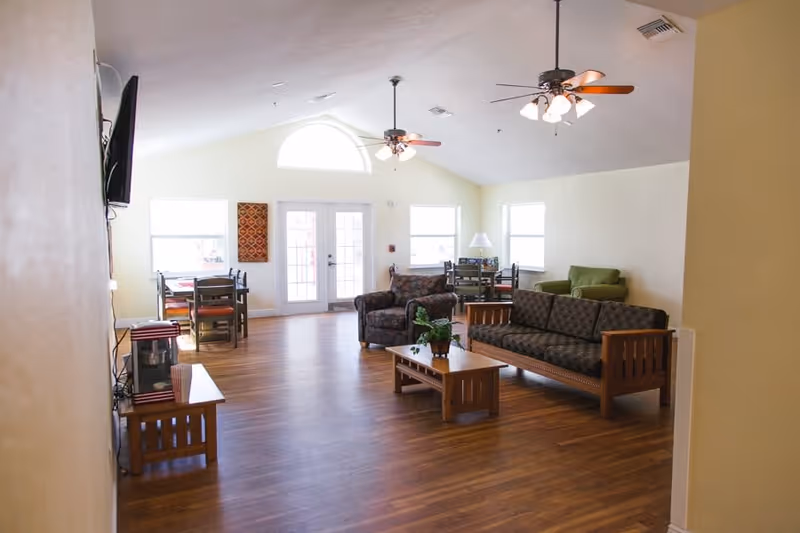 A spacious living room area with wooden flooring, featuring a patterned sofa and armchair, a green armchair, two ceiling fans with lights, a coffee table with a plant, a TV mounted on the wall, and dining tables with chairs near large windows and a door with a half-circle window above it.