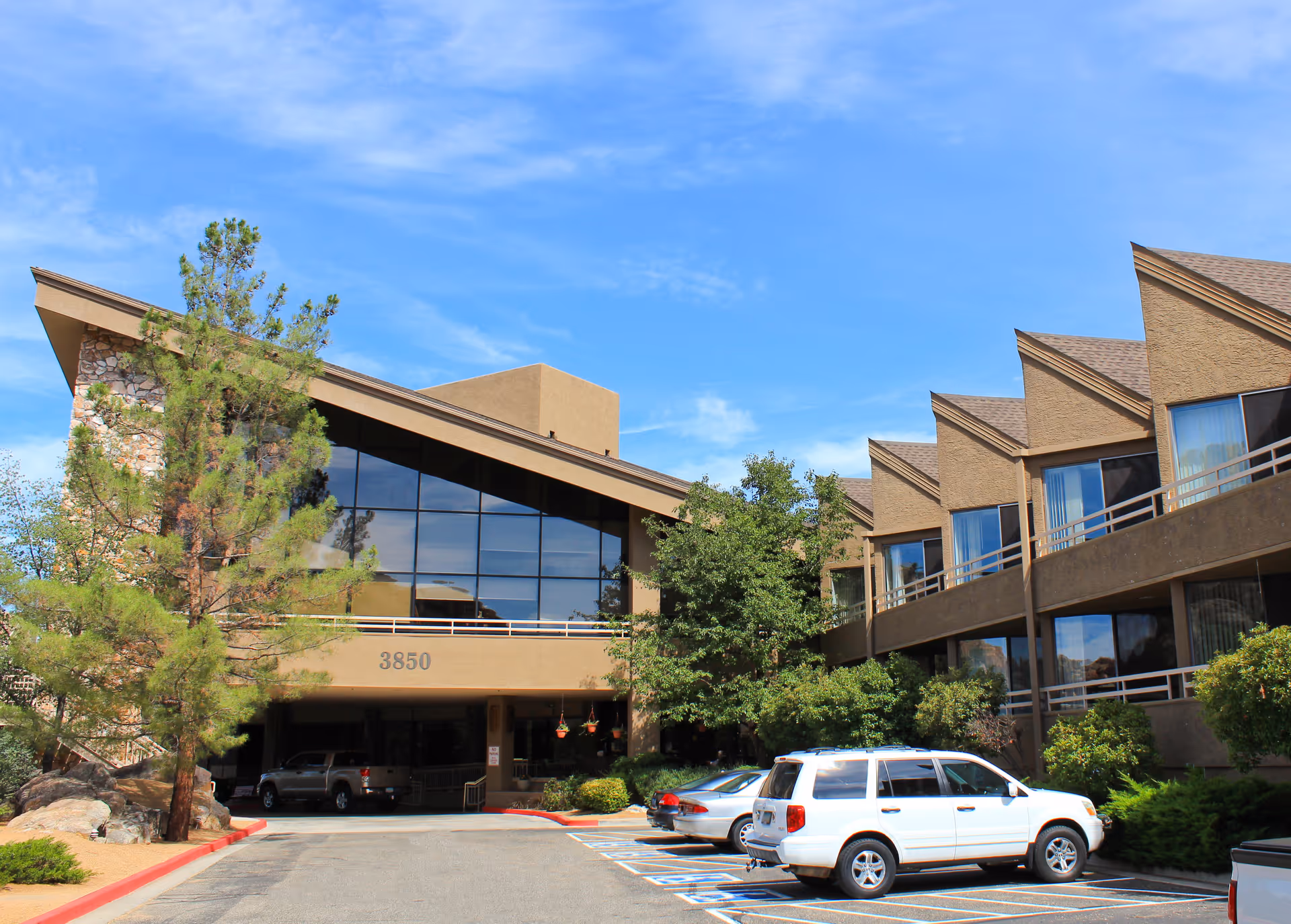 Front exterior of a multi-story senior living building with a large glass entrance, angled rooflines, trees and cars parked in front.