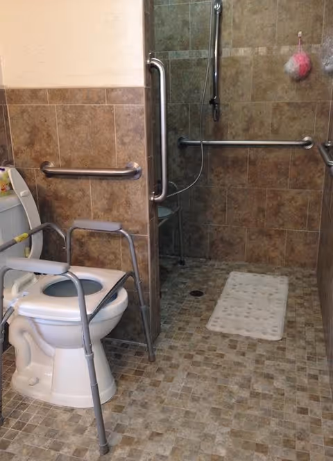 Bathroom with a toilet equipped with a raised seat and armrests for accessibility, grab bars on the walls, and a walk-in shower area with a handheld showerhead and a white bath mat on the tiled floor.
