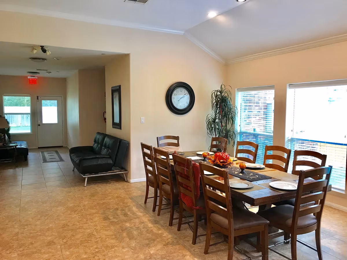 Interior view of a senior living facility dining area with a long wooden table set with plates and autumn-themed centerpiece. The room has large windows with blinds, a wall clock, and a potted plant. Adjacent to the dining area is a hallway with a black leather couch and a door with a window leading outside.