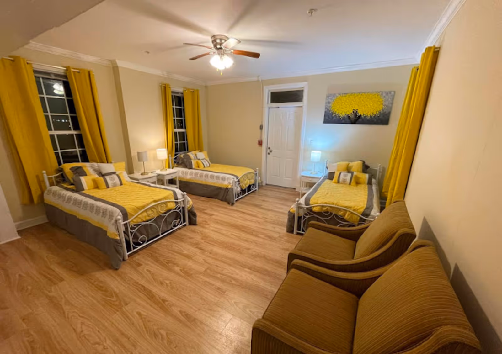 Well-lit shared bedroom with three twin beds dressed in yellow bedding, matching curtains, two armchairs and wood flooring.