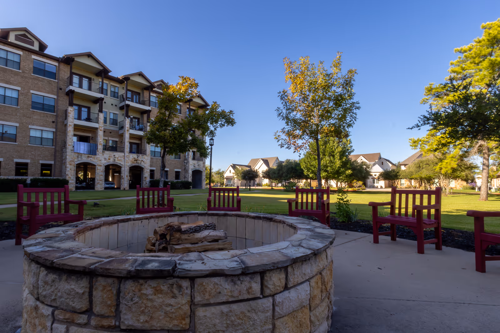 Outdoor seating area with red wooden chairs arranged around a stone fire pit, set on a concrete patio. In the background, there is a multi-story brick building with balconies and a well-maintained lawn with trees and houses under a clear blue sky.