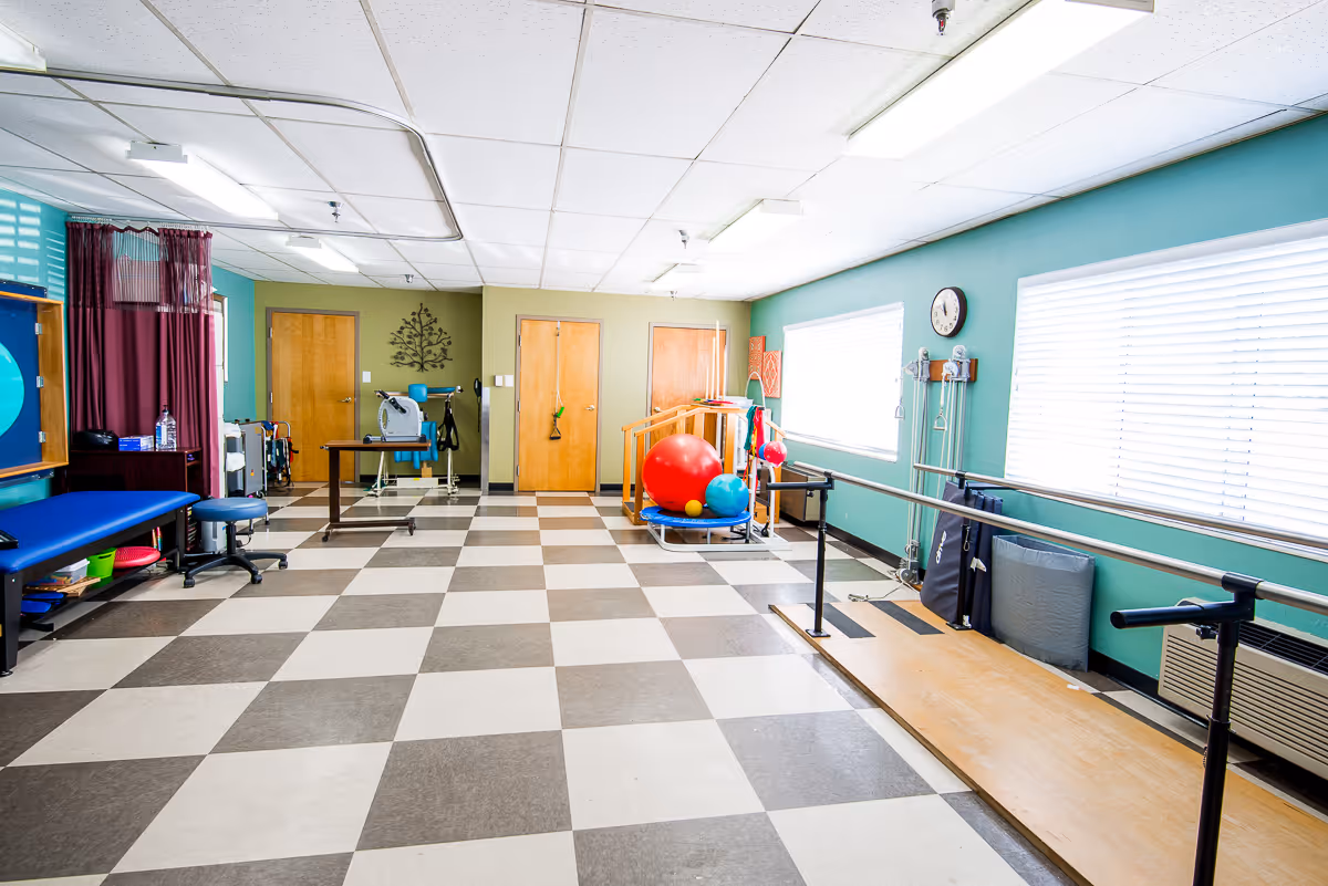 A bright rehabilitation room with checkered tile flooring, teal walls, and large windows with blinds. The room contains physical therapy equipment including parallel bars, exercise balls, a treatment table, and various therapy tools. There are wooden doors and a clock on the wall.