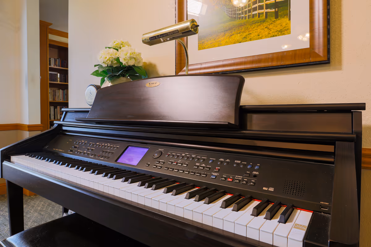 Electric piano in a furnished interior space with a music lamp, framed artwork, and flowers on top.