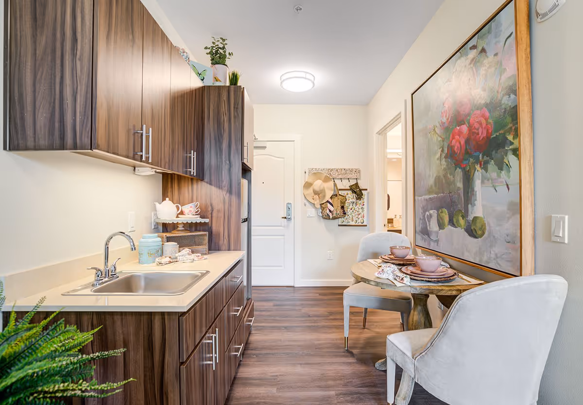 A compact kitchen and dining area in a senior living facility. The kitchen features dark wood cabinets, a stainless steel sink, and a countertop with decorative items. Adjacent to the kitchen is a small round dining table set for two with pink bowls and plates. A large painting of red flowers in a vase hangs on the wall next to the dining table. The floor is wood, and the entrance door is visible in the background with a hat and bags hanging on a wall rack.
