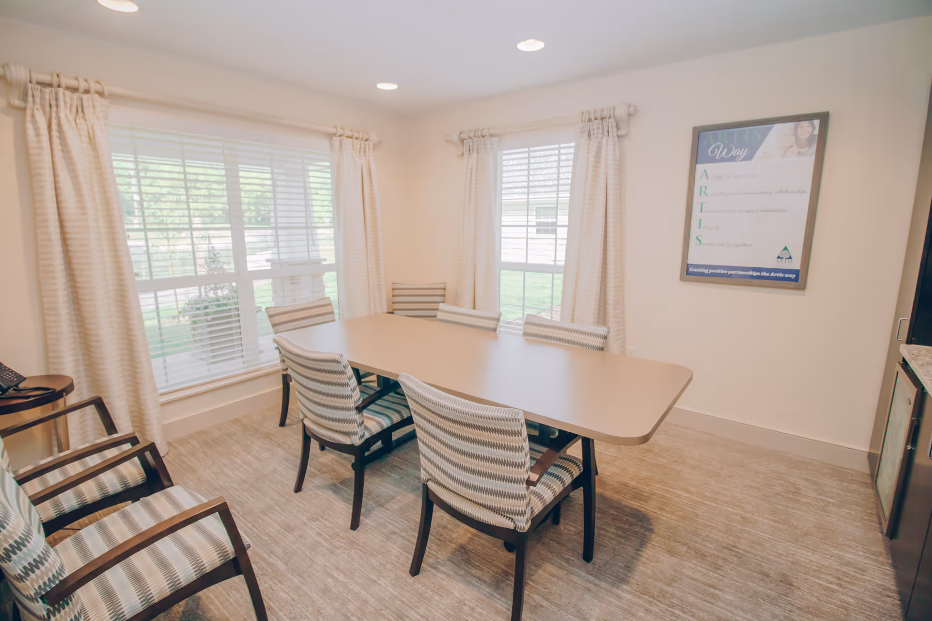 A bright meeting or dining room with a rectangular table surrounded by six striped cushioned chairs. The room has large windows with white curtains allowing natural light to fill the space. A framed poster is visible on the wall, and the floor is covered with a light-colored carpet.