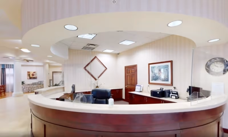 Reception desk area in an assisted living facility with a curved wooden counter, office chair, computer equipment, a hand sanitizer bottle, and framed artwork on the walls. The ceiling has recessed lighting and the space appears clean and welcoming.