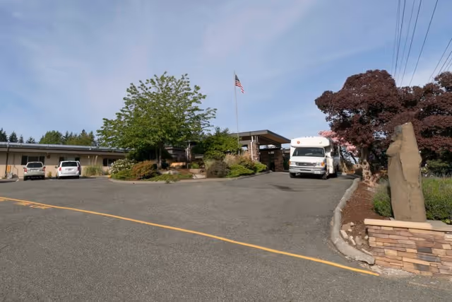Exterior view of Lynnwood Post Acute Rehabilitation Center showing a driveway with parked vehicles, a flagpole with an American flag, landscaped greenery, and a building entrance under a clear sky.