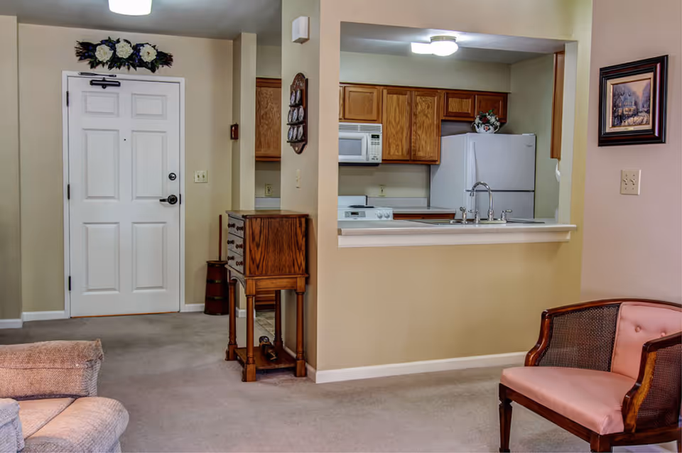 Interior view of a senior living facility showing a small kitchen with wooden cabinets, a white refrigerator, microwave, and stove. In the foreground, there is a pink cushioned chair and part of a beige sofa. A white entrance door is visible on the left side with a decorative floral arrangement above it. The walls are painted beige and there is a small wooden cabinet against the wall near the kitchen.