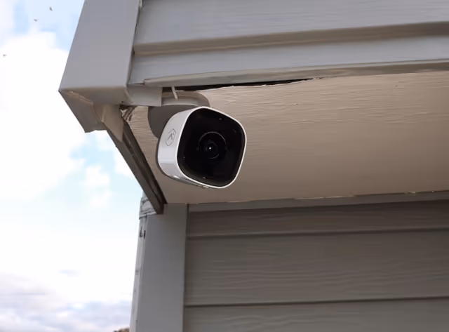 A white security camera mounted under the eaves of a building with gray siding, capturing the outdoor area under a partly cloudy sky.