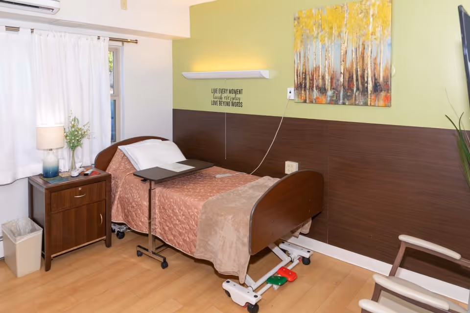 A tidy senior facility bedroom with a hospital-style bed, over-bed table, nightstand with a lamp and flowers, and wall art.