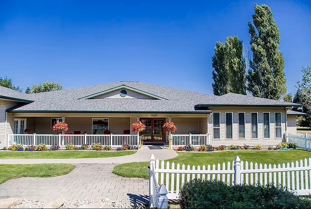 Front exterior of a single-story assisted living building with a covered porch, hanging flower baskets, a white picket fence, and a paved walkway.