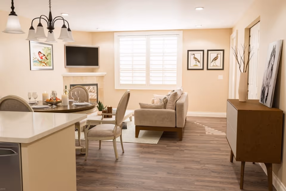 Open-concept living area with a dining table, upholstered sofa, fireplace, wall-mounted TV, and a window with shutters.