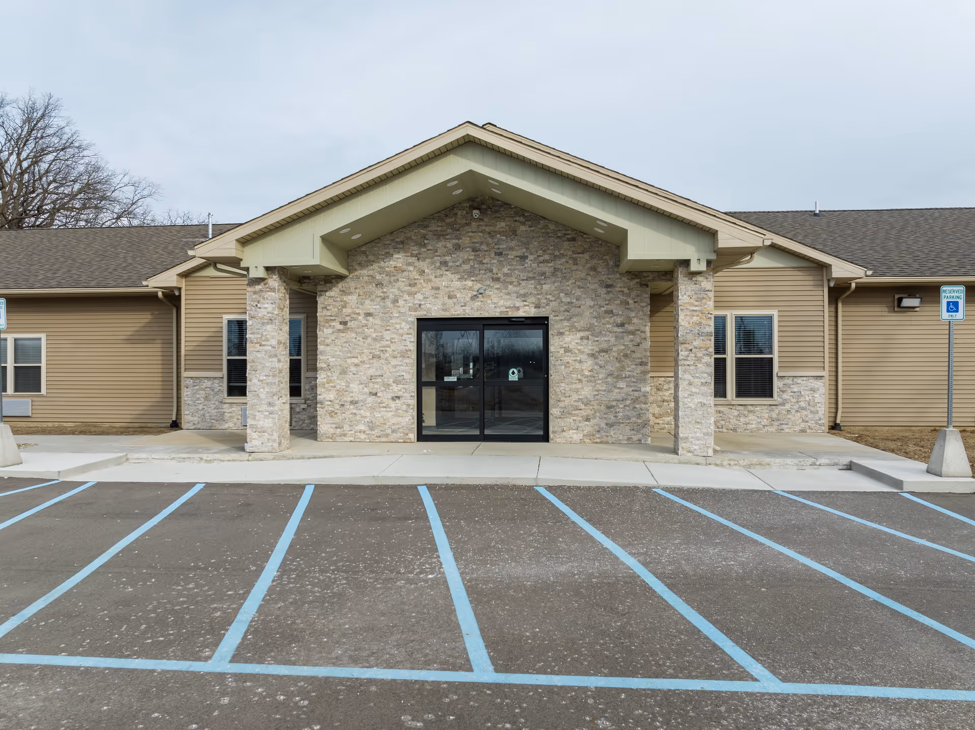 Front exterior view of a single-story building with stone and beige siding, featuring a covered entrance with two stone pillars and glass double doors. There are reserved parking spaces marked with blue lines and handicap signs in front of the building.